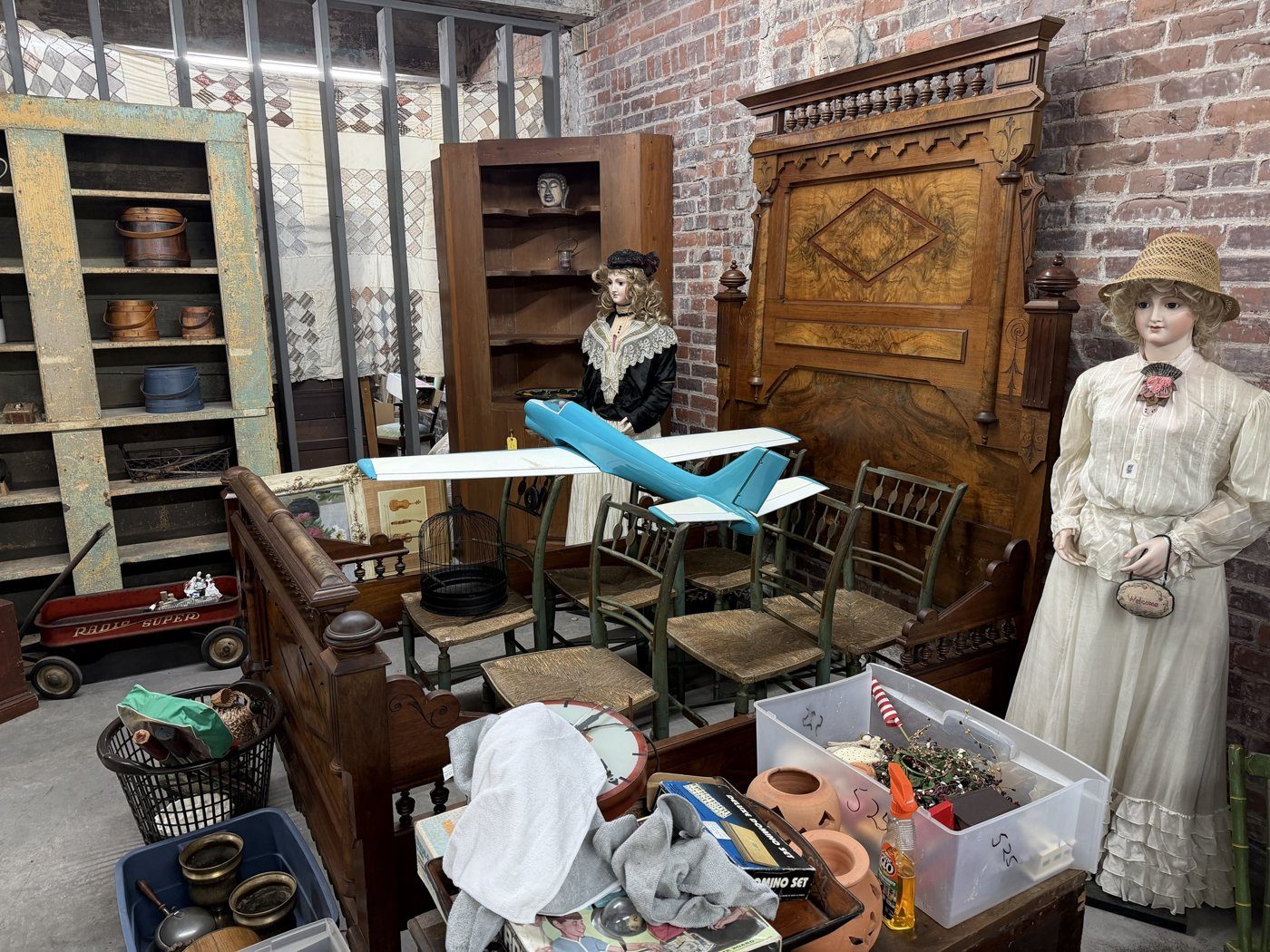 A carved Victorian bed, rush-seat chairs and two dressed mannequins in a brick-walled corner of the shop